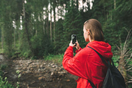 Young tourist woman taking photos on her smartphone in the mountain forest near the river. Back view...