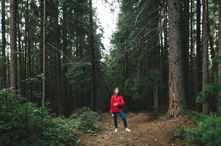 Minimalistic full-length photo of hiker girl in a red jacket in the mountain fir forest. A female...