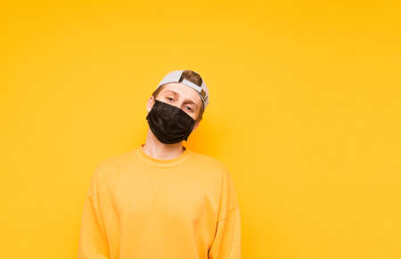 The guy in a cap and a black medical mask stands on a yellow background and poses for the camera with a serious face. Portrait of a young man in a protective gauze mask. Isolated. Coronavirus pandemicの写真素材