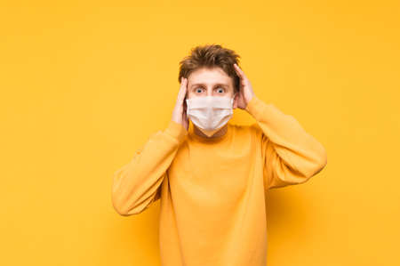 Portrait of a shocked young man in a medical mask on a yellow background, emotionally looking into the camera and holding his head in surprise.Coronavirus pandemic. Quarantine covid-19.の写真素材