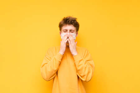 Sick guy in a medical mask sneezes on a yellow background. Young man with a lifeless man wears a...