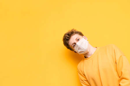 Funny guy in bright casual clothes and medical mask on his face looks from the right side with smile on his face and looks at the camera on a yellow background, isolated.Coronavirus pandemic.COVID-19の写真素材