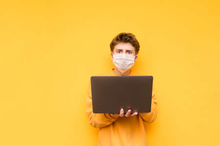 Surprised young man in a medical mask stands with a laptop in his hands and looks at the camera with a shocked face on a yellow background. Coronavirus pandemic. COVID-19. Quarantineの写真素材