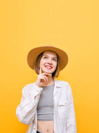 Happy young girl in light summer clothes and hat with thoughtful look looks up at copy space and smiles. Portrait of pensive lady on yellow background, looking up and thinking. Isolated.の写真素材