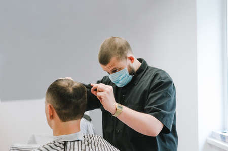 Professional barber in a medical mask cuts the client's hair in a men's barbershop. Work in hairdressers in quarantine. COVID-19の写真素材
