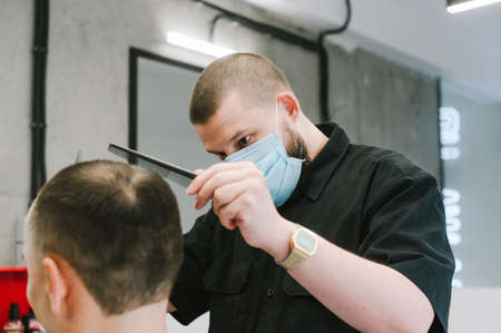 Close photo of a barber in a medical mask cuts a client in a men's barbershop. Barbre cuts the client during quarantine, wears a protective mask. Coronavirus.の写真素材