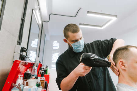 Man hairdresser in a medical mask and with a hair dryer in his hands makes a haircut for a client in a barbershop. Barbershop work during quarantine.の写真素材