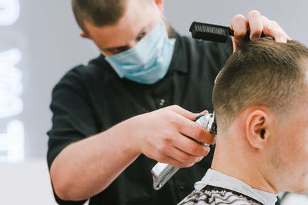 Close photo of the quarantine haircut process, the hairdresser wears a medical mask on his face. Professional barber in a protective mask cuts the client's hair with a clipper.の写真素材