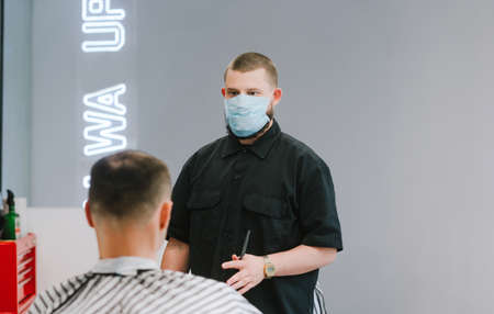 Portrait of a male hairdresser in a medical mask and with scissors in his hands cuts the client in a barbershop. Work of a hairdresser during quarantine, the use of personal protective equipment.の写真素材