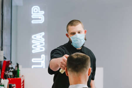 A professional hairdresser in a medical mask works in quarantine, cuts the client with a clipper. Portrait of a professional barber cuts a man in a medical mask. Coronavirus and services.の写真素材