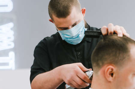 Professional barber in a medical gauze mask on the face cuts the client's hair with a clipper with a focused gaze. Hairdresser works in quarantine. Close upの写真素材