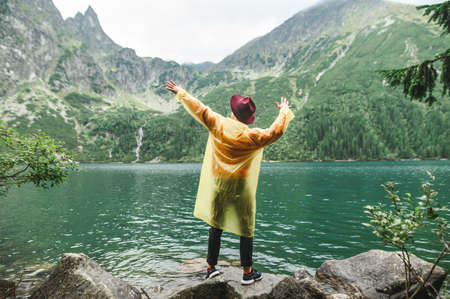 Portrait of a stylish man in a yellow raincoat and hat stands on rocks by the lake overlooking the mountains and shows a Dab. Hipster tourist hiking on a mountain lake, posing for the camera.の写真素材