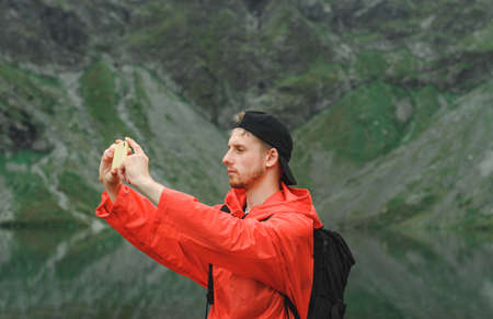 Handsome young tourist in mountains near lake makes nature photo on smartphone. Portrait of a...
