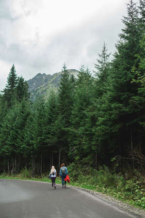 Two female tourists are walking on an asphalt road in the mountains. Hikers hiking, mountain route...