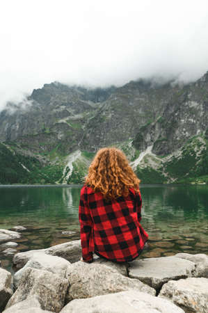 Woman in a red shirt sits on stones in a beautiful lake in the mountains and looks at the landscape. Back of a curly girl with red hair on the background of Lake Morskie Oko in the Tatra Mountainsの写真素材