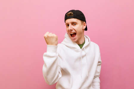 Portrait of joyful guy in white hoodie and cap on pink background, looks into camera and rejoices in...