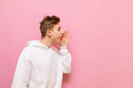 Portrait of a cheerful guy in a white hoodie looks away and shouts with his hand attached. Positive...