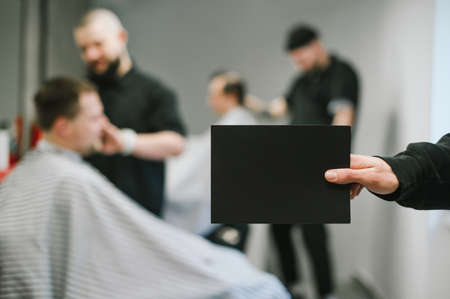 Barber holds a black blank card in his hand for copy space on the background of barbers clipping clients. Male hand holding a blank card on barbershop background. Barbershop advertising, place for textの写真素材