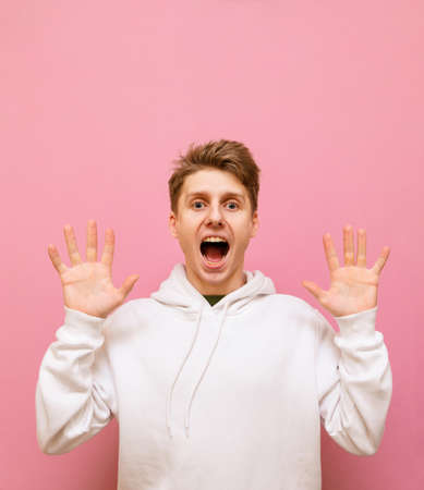 Young frightened man in white hoodie looks into the camera. with shocked face and arms raised up....