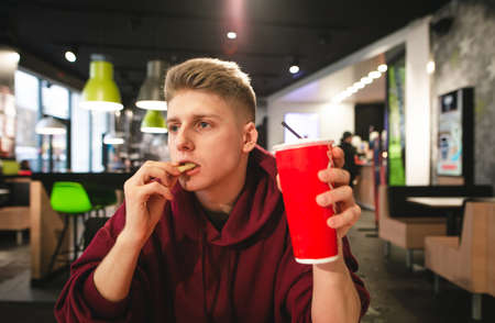 Portrait of a guy in a red hoodie holds a glass of drink in his hands and eats french fries on the...