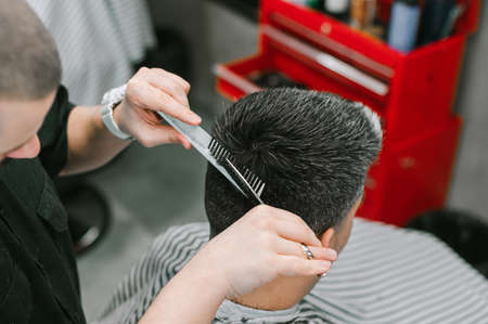 Male hairdresser cuts the hair of an adult client with scissors and a comb. Barber makes a hairstyle...