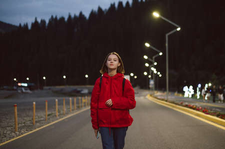 Hiker girl in red raincoat walking on mountain road illuminated by lanterns on coniferous forest background. Attractive girl walking in the evening on an asphalt hiking trail in the mountains.の写真素材