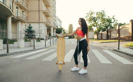 Street girl in stylish summer clothes stands on the street with a longboard in her hands on a sunset...