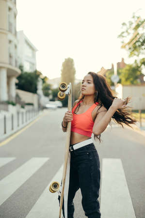 Street girl in stylish clothes stands on the street with a longboard in her hands on a sunset...