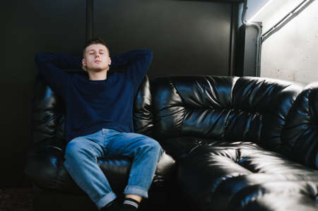 Tired guy in casual casual clothes sits in a dark room on a leather sofa and sleeps. Young man relaxing lying on a black leather sofa in the lounge. COpy spaceの写真素材
