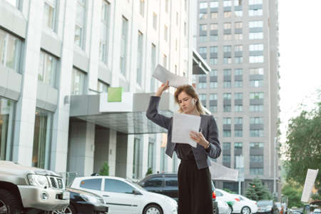 Dissatisfied businesswoman throwing stack of papers in the air in front of the office in the...