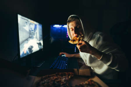 Gamer in white hoodie sits at night at the computer, bites a mouthwatering slice of pizza with his eyes closed and enjoys it. Hungry gamer plays games at home at night and dines with pizza. Fast foodの写真素材