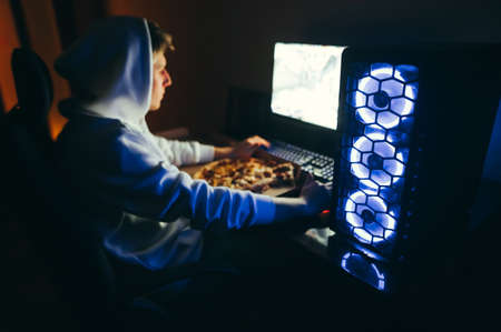 A blue-backed computer stands on a desk against a background of a young male gamer playing video...