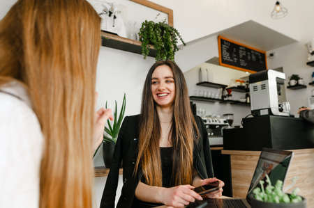 Happy young girlfriends in formal clothes are sitting at a table in a cozy cafe for a break and...