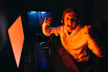 Expressive gamer in headphones and with a microphone plays video games on a computer at night, looks in camera with a crazy face, rejoices to win. Emotional guy is happy to win a computer gameの写真素材
