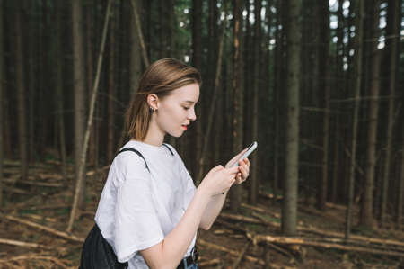Attractive tourist girl in casual clothes and with a ruffle walks through the old dense forest, uses a smartphone and smiles. Pretty hiker girl standing in dark forest with smartphone in hand.の写真素材