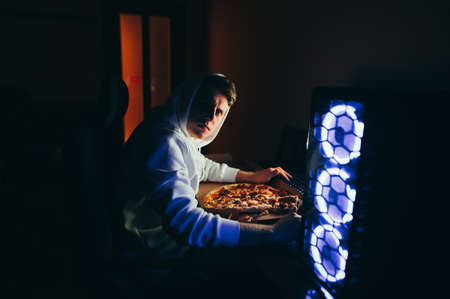 Portrait of a serious young man in a hood sits at home at computer at night, looks into the camera and eats pizza out of the box. Concentrated gamer uses a computer at night and eats fast foodの写真素材