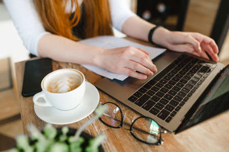 Background Student girl is sitting at a table in a cafe with a cup of coffee, glasses, a laptop and letters in tasks, typing on a keyboard. Copy space.の写真素材