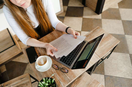 Freelancer girl sits at a coffee table with business papers in her hands and uses the internet on a...