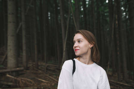 Portrait of a smiling girl in a white T-shirt stands in the thick dark forest, looking away with a smile on her face. Hipster girl hiking in mountain forest posing at camera and smiling.の写真素材