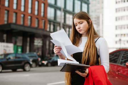 Concentrated girl with long hair standing on the street, background of a cityscape examining...