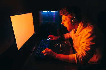 Portrait of emotional gamer in headphones playing computer games at home at table at night, yelling into microphone and looking intently at computer screen. Expressive gamer broadcasts a game streamの写真素材