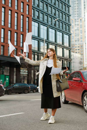Girl with long hair stands on the street and throws up notes with a serious face, vertical photo. Young girl student throws away paper from anger on cityscape background.の写真素材