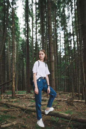 Pretty hipster girl in stylish casual casual hiking, standing in dense coniferous forest and looking away with a serious face. Vertical portrait.の写真素材
