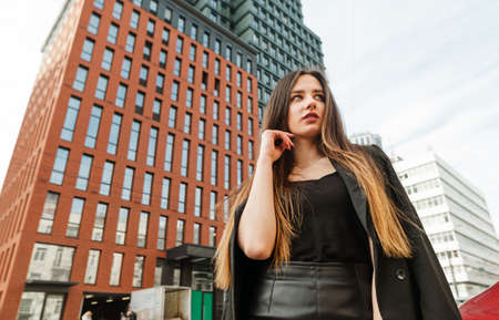 Stylish young business woman in dark casual clothes posing at camera with serious face on skyscraper...