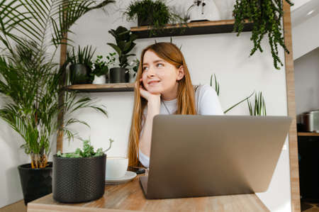 Smiling lady sits in a cozy cafe with flowerpots, uses a laptop and looks away with a smile on her...
