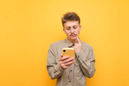 Thoughtful young man with mustache and glasses uses a smartphone against a yellow background, looks at the screen and thinks, wearing a shirt.Funny nerd with a serious face is using the phone. Isolatedの写真素材