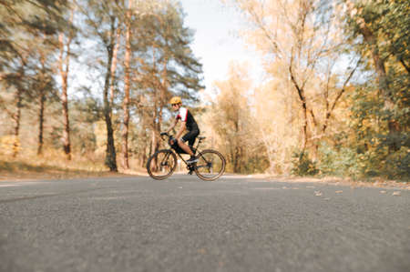 Background. Blurred photo, focus on asphalt. Asphalt road in the woods and a cyclist walking on a bicycle in sports gear. Cyclist rides through the city in the autumn forest.の写真素材