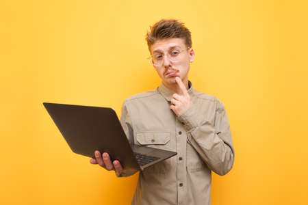 Funny young man freelancer isolated on yellow background with laptop in hand looking into camera with pensive face. Nerd in shirt and glasses with laptop in hand stands on yellow background and worksの写真素材