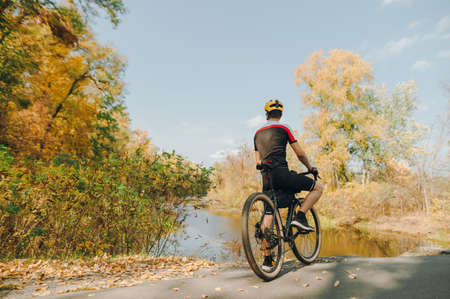 Professional cyclist in helmet and sportswear stands with bicycle, background of lake and autumn...