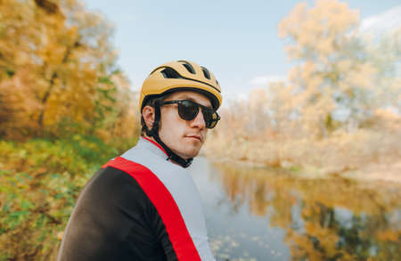 Closeup portrait of handsome cyclist man in sunglasses and helmet on background of beautiful autumn park landscape, looking into camera with serious face. Serious cyclist posing on camera outdoors.の写真素材